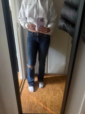 Mörkblå bootcut jeans med slitning - Säljer ett par mörkblå bootcut jeans med slitning vid ena knät. Jeansen har klassisk femficksdesign, normal midja och är tillverkade i stretchigt denimtyg för skön passform. Perfekta för en avslappnad och trendig look.