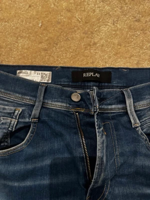 Blå jeans från Replay - Säljer ett par klassiska blå jeans från Replay med snygga slitningar och kontrastsömmar. Jeansen har fem fickor, dragkedjegylf och läderpatch med logga bak i midjan. Perfekta för en avslappnad och trendig look.