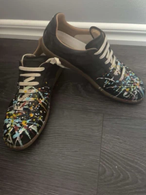 Maison margiela paint splatter gats - Unika svarta sneakers från Maison Margiela med coola färgstänk i blått, gult, vitt och rött över hela ovandelen. Skorna har beige snörning och bruna sulor. Perfekt för dig som vill sticka ut med en kreativ och trendig look.