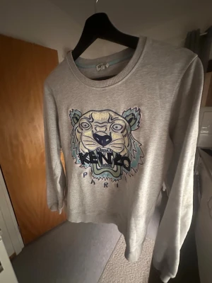 Grå Kenzo sweatshirt med tiger - Cool grå sweatshirt från Kenzo med ikoniskt tigerbroderi i blått, gult och lila på bröstet. Tröjan har rund halsringning och långa ärmar. Perfekt för dig som vill sticka ut med ett trendigt plagg från ett välkänt märke.