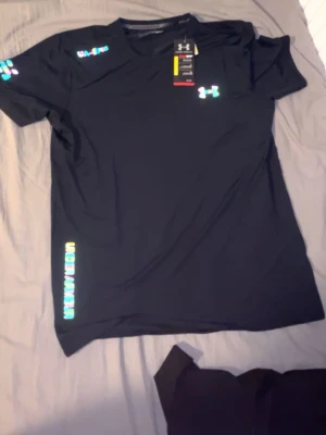 Svart träningsset från Under Armour - Säljer ett svart träningsset från Under Armour med t-shirt och shorts. Båda har reflekterande loggor och detaljer i neonfärger. Materialet är lätt och snabbtorkande, perfekt för gym eller löpning. Shorts med elastisk midja och snörning.