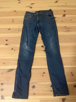 Blå slim fit jeans från Replay - Snygga blå slim fit jeans från Replay med klassiska fem fickor och coola slitningar på både fram- och baksidan. Jeansen har en läderpatch med Replay-logga på bakfickan och kontrastsömmar. Perfekta för dig som gillar en avslappnad och trendig look. Men som man ser har den några vita prickar där bak kan säkert gå att fixa på något sätt men annars bra skick