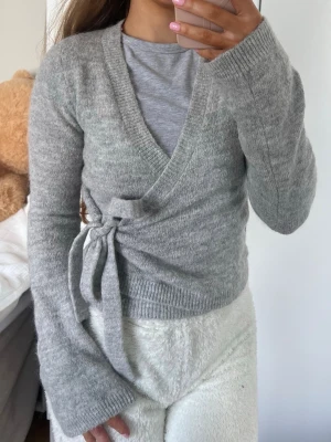 Kofta - Mysig grå kortare omlottkofta med långa ärmar och knytband i sidan. Koftan har ribbade muddar och en mjuk, stickad känsla som passar perfekt till lager på lager. Enkel att matcha med olika outfits för en avslappnad och trendig look. Storlek XS från Gina Tricot 🩶