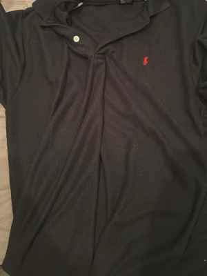 Svart pikétröja från Ralph Lauren - Klassisk svart pikétröja från Ralph Lauren med röd broderad logga på bröstet. Tröjan har korta ärmar, krage och knappar vid halsen. Tillverkad i mjuk bomull som känns skön mot huden. Perfekt för en clean och stilren look.