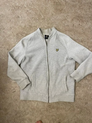 Ljusgrå ziptröja från Lyle & Scott - Ljusgrå ziptröja från Lyle & Scott med klassisk gul logotyp på bröstet. Tröjan har ribbade muddar, två fickor och en diskret struktur i materialet. Perfekt för en avslappnad och stilren look.