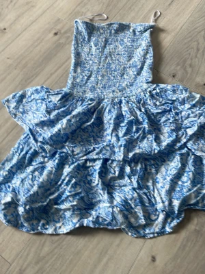 Blåvit sommarklänning från FB Sister - Supersöt blåvit klänning från FB Sister i butiken New Yorker med strapless. Klänningen har ett volangigt, flowy kjolparti och ett blommigt mönster. Perfekt för varma dagar och ger en fräsch och ungdomlig vibe. 