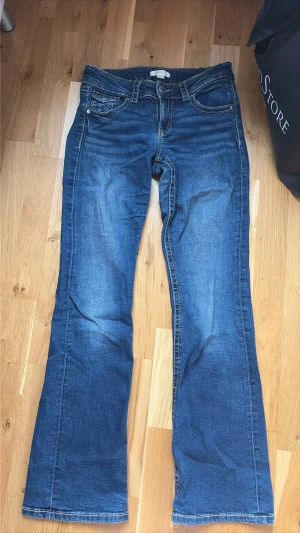 Blå bootcut jeans - Snygga lågmidjade blå bootcut jeans med knappfickor på baksidan. Har även ett litet hål vid foten och även lite sliten, vilket man inte lägger så mycket märke till.