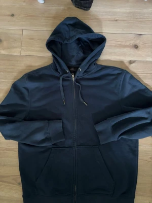 Mörkblå hoodie med dragkedja H&M - Mörkblå hoodie från H&M i regular fit med dragkedja framtill, huva med dragsko och två stora fickor. Tillverkad i mjukt bomullsmaterial som är skönt mot huden. Perfekt för en avslappnad och stilren look.