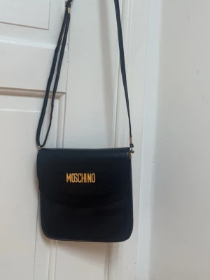 Svart axelväska från Moschino - Snygg svart axelväska från Moschino med justerbar rem och guldfärgad logga framtill. Väskan har en stilren, fyrkantig form och är tillverkad i slätt material som ger en lyxig känsla. Perfekt för dig som gillar statement-accessoarer.
