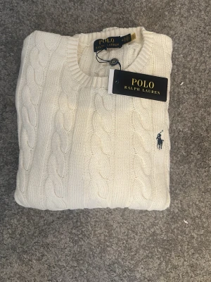 Vit kabelstickad tröja Polo Ralph Lauren - Kabelstickad vit tröja från Polo Ralph Lauren med rund halsringning och broderad logga på bröstet. Klassisk och stilren design i mjukt material, perfekt för lager-på-lager. Storlek M