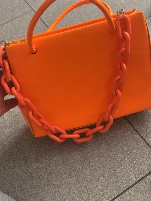 Neonorange handväska med kedja - Cool neonorange handväska i fyrkantig modell med matchande plastkedja och handtag. Väskan har guldfärgade metalldetaljer och ett rymligt huvudfack. Perfekt statement piece för dig som vill sticka ut.