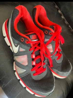 Nike svarta och röda sneakers - Snygga sneakers från Nike i svart och rött med vita detaljer. Skorna har en klassisk låg siluett, röda skosnören och meshpaneler för extra ventilation. Swoosh-logga på sidan och vadderad insida för skön komfort. Perfekta för dig som gillar sportig stil.