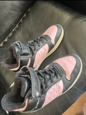 Svarta och rosa high-top sneakers WOO - Snygga high-top sneakers från WOO i svart och rosa med logotypmönster på sidorna. Skorna har snörning, kardborreband upptill och mjukt foder för extra komfort. Perfekta för dig som vill sticka ut med en cool streetstyle-look.