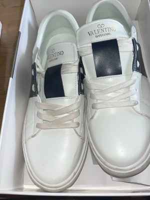 Valentino Garavani vita sneakers - Snygga vita sneakers från Valentino Garavani med breda mörkblåa band över ovandelen och vita snören. Skorna har en klassisk låg siluett och är tillverkade i slätt läder med subtila nitar på sidorna. Perfekta för dig som gillar stilrena och exklusiva detaljer. Bara använd en gång priset kan sänkas privat 