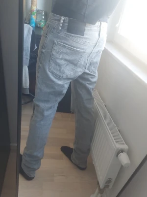 Jack & jones - Säljer ett par grå jeans med raka ben och slitna detaljer på knän och fickor. Jeansen har klassisk femficksdesign och design på bakfickorna
