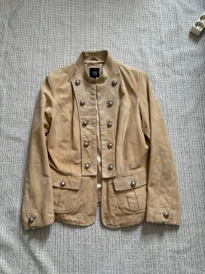 Beige mockakavaj med dekorativa knappar - Stilren beige kavaj i mocka från Wilsons Leather med militärinspirerad design. Kavajen har ståkrage, dubbla rader stora dekorativa metallknappar och två fickor framtill. Materialet är i äkta läder samt mjukt och ger en exklusiv känsla. Perfekt för dig som vill sticka ut med en unik look. Lite oversize på mig som bär XS har en liten fläck på framsidan men går att tvätta bort!