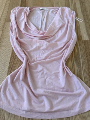 Ljusrosa ärmlös topp med drapering - Säljer en ljusrosa ärmlös topp från H&M med snygg draperad halsringning. Toppen är i mjuk bomull och har en feminin, figurnära passform. Perfekt att styla med jeans eller kjol för en trendig look.