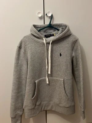 Grå hoodie från Polo Ralph Lauren - Snygg grå hoodie från Polo Ralph Lauren med klassisk huva och vita dragsnören. Har en stor magficka och den ikoniska lilla broderade loggan på bröstet. Perfekt för en chill och avslappnad stil. Mjuk och skön bomullsblandning. Endast använd några gånger inget man märker. Storlek M men passar S så om due runt 170-180 så kommer den här passa dig perfekt. Kom gärna med pris förslag 