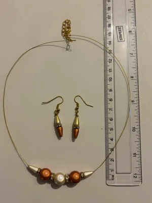 Smyckesset med pärlor i guld och brun - Stilrent halsband med tunnt guldigt band och tre pärlor i brunt och vitt, samt guldfärgade detaljer. Matchande örhängen med konformade pärlor i vitt och brunt. Perfekt accessoar för att lyfta din outfit. Nickelfria