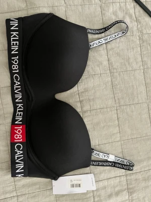 Svart Calvin Klein 1981 bh - Svart vadderad bh från Calvin Klein med bred resår och logga 'CALVIN KLEIN 1981' i vitt och rött. Axelbanden är justerbara och har samma logga. Snygg och sportig design, perfekt för dig som gillar stilrena underkläder.