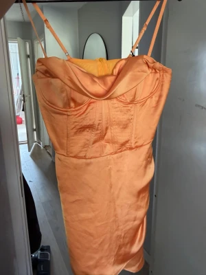 Orange satin kortklänning med smala band - Snygg orange kortklänning i satin med smala axelband och draperad urringning. Klänningen har figurnära passform och dragkedja i ryggen. Perfekt för dig som vill sticka ut med en färgstark look.
