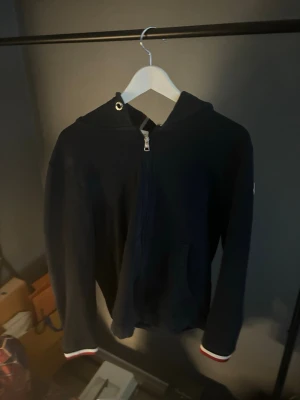 Svart Moncler hoodie med dragkedja - Svart hoodie från Moncler med dragkedja framtill, huva och fickor. Ärmsluten har rödvita detaljer och Moncler-logga sitter på ena ärmen. Huvan har en snygg rand i rött och vitt. Perfekt för en stilren och sportig look.