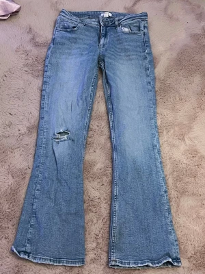 Ljusblå bootcut jeans med slitning - Säljer ett par ljusblå bootcut jeans med klassisk femficksdesign och en snygg slitning på ena benet. Jeansen har utsvängda ben och är tillverkade i denim. Perfekta för dig som gillar en avslappnad och trendig stil.