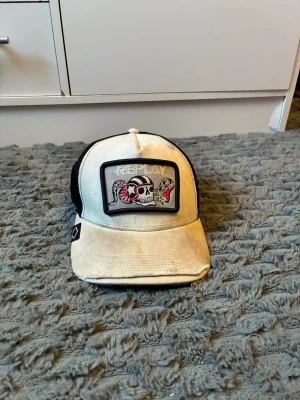Vit och svart keps från Replay - Säljer en vit och svart keps från Replay med ett stort tygmärke framtill som har en döskalle och ormar i rött och svart. Kepsen har mesh på baksidan och justerbar snapback. Skön streetstil och cool detalj till din outfit.