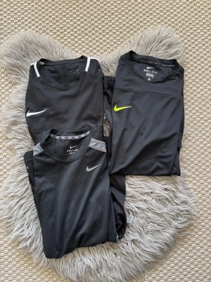 3-Pack Nike Dri-Fit svarta tränings-tshirts - Säljer flera svarta Nike Dri-Fit tränings-tshirts i funktionsmaterial. T-shirtsen har olika detaljer som vita, grå eller blå inslag samt Nike-logga i vitt eller gult. Perfekta för träning med sin lätta och snabbtorkande kvalitet.