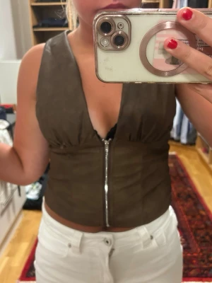 Brun mocka korsett med dragkedja - Snygg brun korsettopp i mockaimitation med djup v-ringning och dragkedja framtill. Toppen är ärmlös och har en croppad passform som ger en trendig look. Perfekt att styla med jeans eller kjol för en cool outfit.