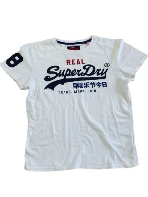 Vit Superdry t-shirt med tryck - Vit t-shirt från Superdry med stort blått och rött tryck framtill och siffra på ärmen. Klassisk rund halsringning och korta ärmar. Tillverkad i mjuk bomull för en skön känsla.