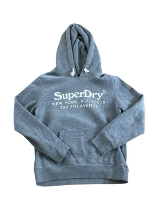 Grå hoodie från Superdry - Snygg grå hoodie från Superdry med stor vit logga och text på bröstet. Klassisk huva med dragsko och magficka framtill. Tillverkad i mjukt sweatshirtmaterial som är perfekt för chill dagar. Ribbade muddar vid ärmslut och nederkant.