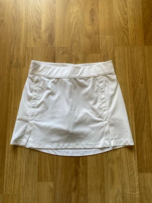 Vit tenniskjol från J.Lindeberg - Snygg vit tenniskjol från J.Lindeberg med bred midjeresår och diskreta sömdetaljer. Kjolen är tillverkad i ett lätt och stretchigt material som passar perfekt för träning eller golf. Enkel och stilren design.