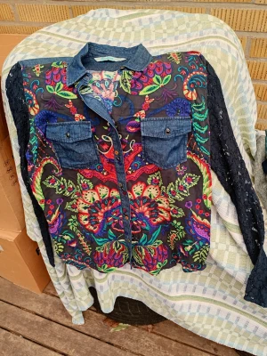 Blommig skjorta med jeansdetaljer xxl (M/L - Färgstark skjorta med blommigt mönster i rött, grönt, blått och lila. Skjortan har jeanskrage, jeansfickor och knappar framtill samt svarta spetsärmar. Perfekt för dig som gillar unika plagg med mycket färg och detaljer.