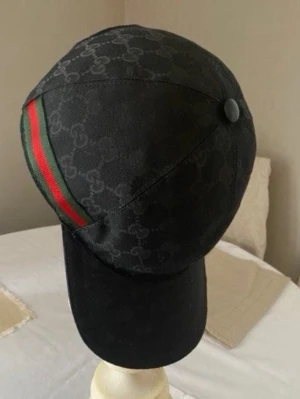 Svart keps med mönster - Snygg svart keps med det klassiska mönstret över hela kepsen och en grön-röd rand på sidan. Kepsen har böjd skärm och knapp på toppen. Perfekt accessoar för dig som vill ha en exklusiv och trendig look.