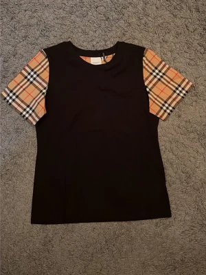 Burberry t-shirt  - Säljer en  svart t-shirt från Burberry med rutiga ärmar i beige, svart, vit. den är tillverkad i mjukt material som känns skönt mot huden. Perfekt för dig som gillar stilrena designer. L passar M