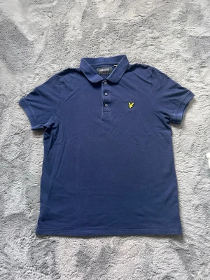 Mörkblå pikétröja från Lyle & Scott - Klassisk mörkblå pikétröja från Lyle & Scott med korta ärmar och gul logga broderad på bröstet. Tröjan har krage och knäppning med tre knappar framtill. Tillverkad i mjuk bomull som känns skön mot huden.