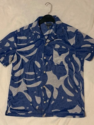 Blå mönstrad skjorta från H&M - Säljer en blå skjorta från H&M med abstrakt mönster i vit. Skjortan har korta ärmar, klassisk krage och knappar framtill. Passar perfekt för varma sommardagar.