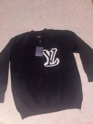 Svart stickad tröja - Snygg svart stickad tröja med stor vit logga på bröstet. Tröjan har rund halsringning och ribbade muddar vid ärmslut och nederkant. Perfekt för dig som gillar exklusiva och trendiga plagg.