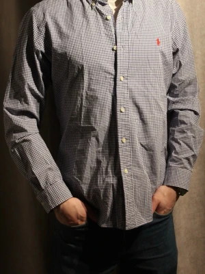 Blårutig skjorta från Polo Ralph Lauren - Säljer en klassisk blårutig skjorta från Polo Ralph Lauren med vit och blå små rutor och ikonisk röd logga på bröstet. Skjortan har knappar framtill, krage och långärmad passform. Perfekt för dig som gillar stilrena och tidlösa plagg.