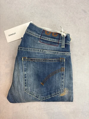 Blå slim fit jeans från Dondup - Snygga blå slim fit jeans från Dondup med slitna detaljer och lätt tvättad look. Klassisk femficksmodell med kontrastsömmar och läderpatch bak i midjan. Perfekta för en avslappnad och trendig stil.