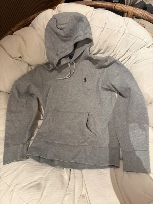 Grå hoodie från Polo Ralph Lauren - Snygg grå hoodie från Polo Ralph Lauren med snygg design. Har en broderad blå logga på bröstet och en stor magficka. Tillverkad i mjukt bomullsmaterial. Perfekt för en avslappnad och stilren look. Den är helt i nyskick har bara använt den två gånger, säljer den då jag inte använder den😊