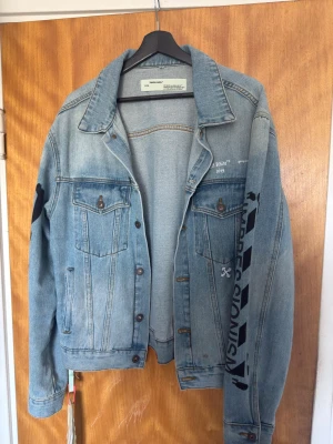 Off-White ljusblå jeansjacka - Nypris 8000kr. Snygg ljusblå jeansjacka från Off-White med ikoniska svarta tryck på ryggen och ärmen. Klassisk modell med bröstfickor och knappar framtill. Jackan har en avslappnad passform och coola detaljer som gör den unik.