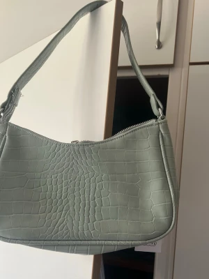 Grön axelväska i krokopräglat mönster - Snygg ljusgrön handväska med axelrem och krokopräglat mönster. Väskan har en mjuk, rundad form och stängs med dragkedja upptill. Perfekt accessoar för att lyfta din outfit med trendig struktur och färg.