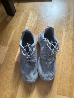 Grå Nike Roshe One sneakers - Snygga grå Nike Roshe One sneakers med vit sula och vit swoosh på sidan. Ovandelen är i meshmaterial som andas och skorna har klassisk snörning. Perfekta för en sportig och avslappnad stil.