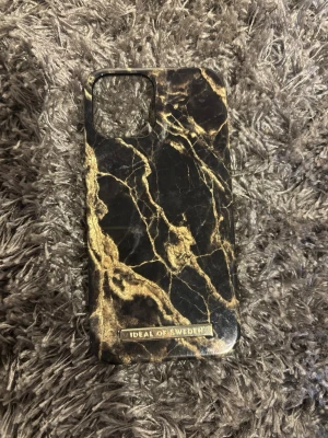 Svart marmormönstrat mobilskal iDeal iPhone 12 - Snyggt mobilskal från iDeal of Sweden med svart bakgrund och guldiga marmordetaljer. Skalets yta är slät och hård, tillverkat i plast. Passar perfekt för dig som vill ha ett stilrent och trendigt skydd till din mobil. Pris kan diskuteras 