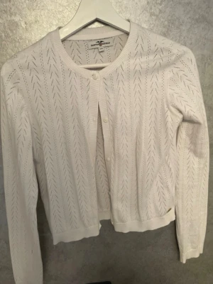 Vit stickad kofta Hampton replublic - Snygg vit stickad kofta från Hampton replublic med diskret hålmönster och rund halsringning. Koftan har långa ärmar, ribbade muddar och knäppning framtill med små knappar. Perfekt att slänga över en topp för en fräsch look.