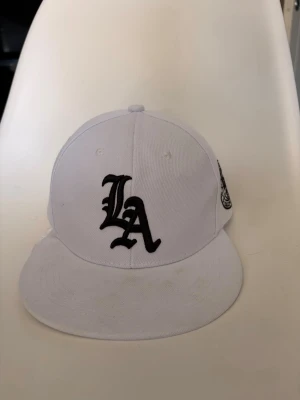 Vit LA keps med broderad logga - Säljer en vit keps med svart broderad LA-logga framtill och en rund Los Angeles-brodyr på sidan. Klassisk snapback-modell med platt skärm och justerbar baksida. Tillverkad i bomullstyg, perfekt för en streetwear-look.