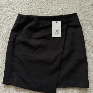 Svart omlottkjol Bikbok stl 34 - Stilren svart kortkjol från Bikbok i omlottmodell. Kjolen har en rak passform och stängs med dragkedja bak. Perfekt för dig som gillar minimalistisk och clean stil. Materialet känns mjukt och följsamt.