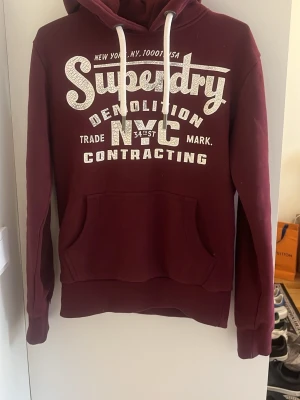 Vinröd hoodie från Superdry - Snygg vinröd hoodie från Superdry med vit tryckt logga och text på bröstet. Klassisk känguruficka framtill, huva med vita snören och ribbade muddar. Perfekt för en avslappnad och trendig stil.